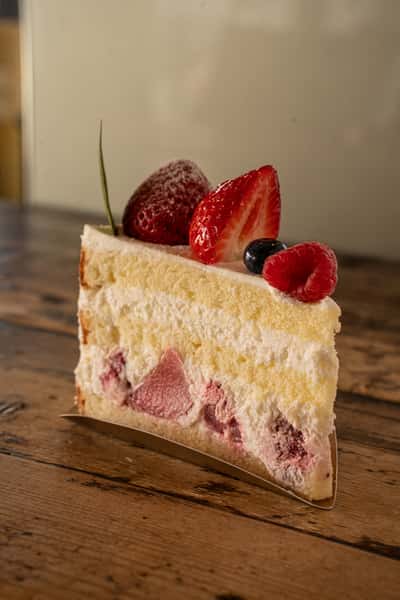 A slice of fluffy strawberry cream cake topped with fresh strawberries, raspberry, and blueberry, served on a rustic wooden table.