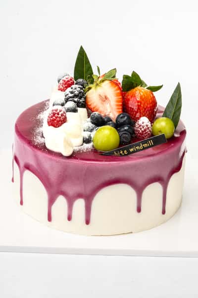 Berry drip cake with glossy purple glaze, topped with fresh strawberries, blueberries, raspberries, green grapes, and whipped cream, styled as an elegant artisan bakery dessert.