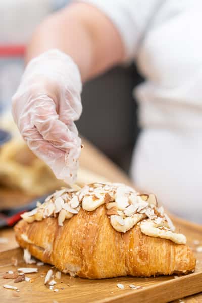 Pastry chef sprinkling sliced almonds over a freshly baked almond croissant on a wooden board, showcasing artisanal bakery preparation and flaky pastry texture.