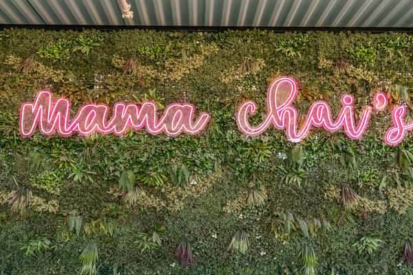 Neon pink "Mama Chai's" sign mounted on a lush green vertical garden wall, blending modern branding with a nature-inspired ambiance.