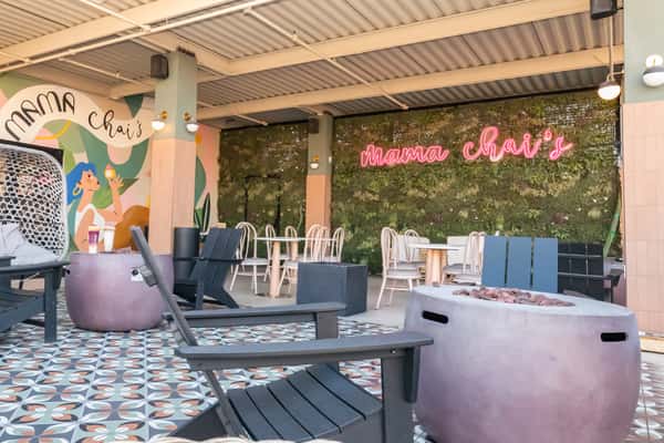 Outdoor patio seating at Mama Chai's with black lounge chairs, round fire pit, modern tiles, and neon pink signage set against a vertical garden—ideal for cozy gatherings and stylish boba tea hangouts.
