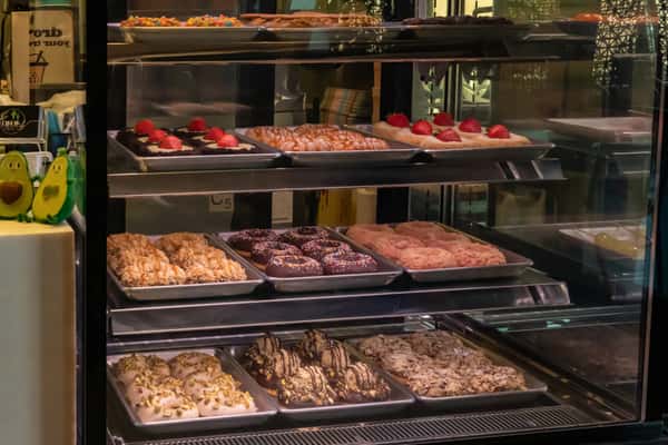 Glass bakery display case filled with assorted gourmet donuts and pastries, including chocolate sprinkle donuts, strawberry-topped treats, and nut-covered confections, creating a colorful and indulgent presentation.
