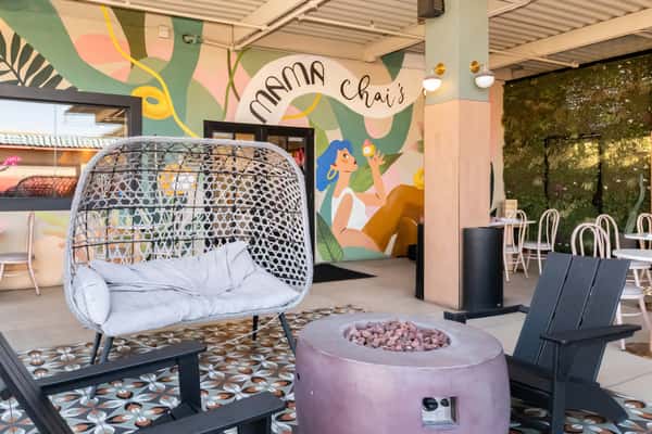 Whimsical patio corner at Mama Chai's featuring a woven hanging chair, a modern fire pit, and a colorful mural of a woman enjoying dessert, enhancing the café's playful and Instagrammable ambiance.