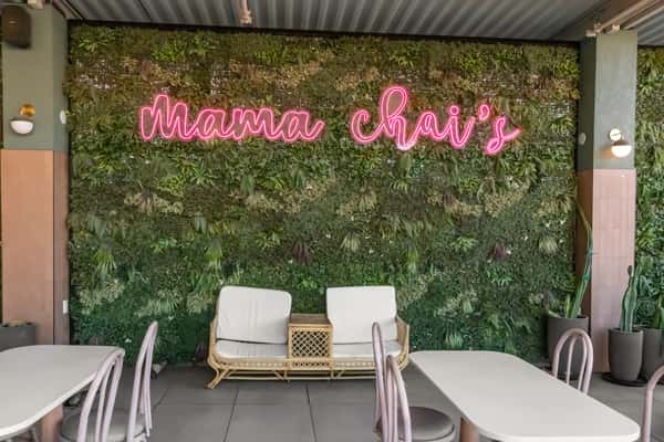 Cozy outdoor seating area at Mama Chai's featuring modern rattan furniture and a lush green plant wall with pink neon signage.