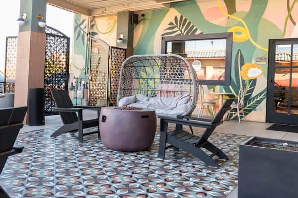 Chic outdoor seating area with patterned tile flooring, a wicker egg chair, and bold tropical wall murals, creating a cozy, Instagram-worthy café vibe.