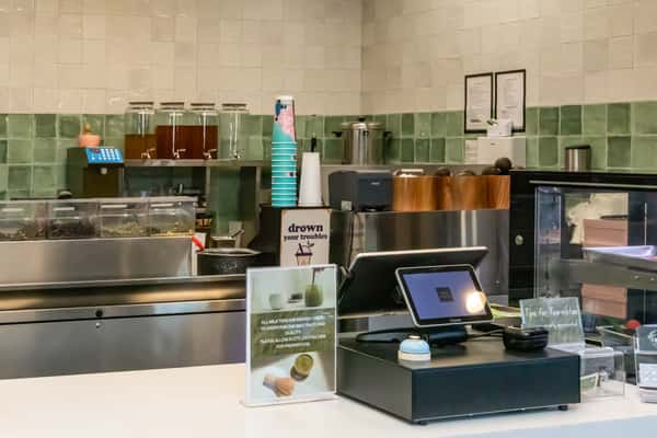 Close-up of tea shop counter with clear tea canisters, pastel paper cups, and signs promoting matcha drinks and gluten-free options.