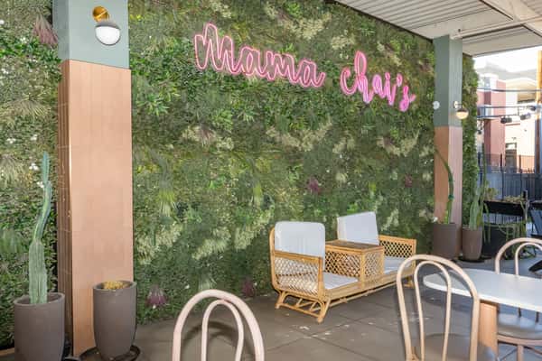 Trendy outdoor seating area at Mama Chai's, featuring modern wicker lounge chairs with white cushions, a wooden table, and a lush green plant wall with a neon pink sign. The chic café patio is accented with potted cacti and natural light, creating a vibrant, Instagram-worthy atmosphere ideal for casual dining or sipping bubble tea.