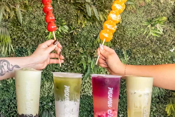 Colorful display of Mama Chai's signature bubble teas and glossy candied fruit skewers, held in front of a vibrant green plant wall. The drinks include matcha latte with boba, layered matcha-taro, hibiscus lemonade, and creamy golden milk tea, paired with bright red candied strawberries and glazed mandarin skewers—perfect for aesthetic café content and Asian-inspired dessert lovers.