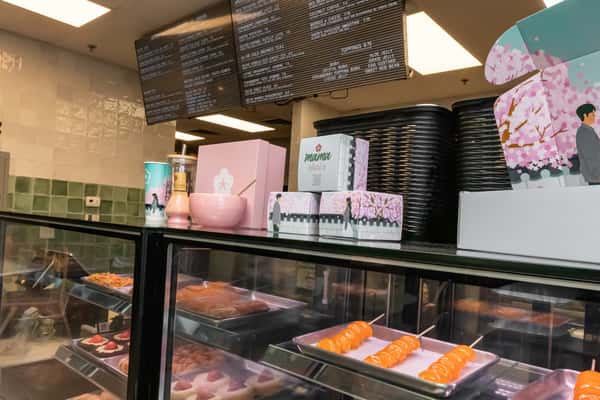 Mama Chai's counter featuring pastel pink cherry blossom-themed dessert boxes, skewered glazed treats, and a sleek digital menu. Aesthetic interior with takeout-ready sweets and signature packaging.