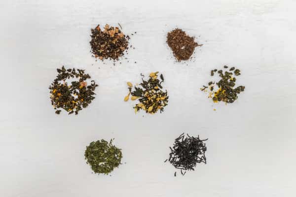Assorted loose leaf teas arranged in small piles on a white surface, including blends of green, black, and herbal teas with visible flower petals, fruit pieces, and spices.