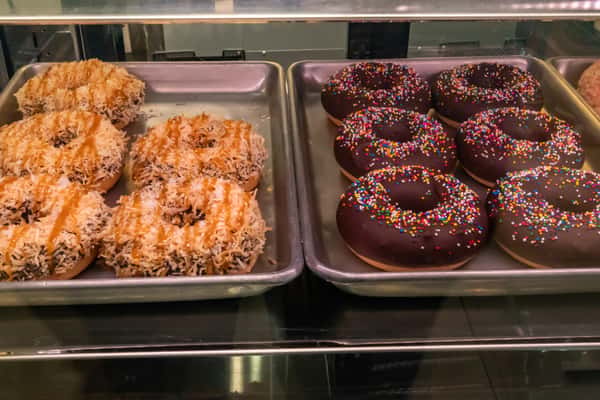 Freshly baked donuts on display; left tray features coconut caramel donuts topped with toasted shredded coconut and caramel drizzle, while the right tray showcases classic chocolate glazed donuts with rainbow sprinkles.