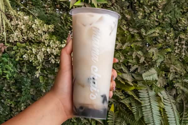 Hand holding an iced milk tea with black boba pearls in a clear Mama Chai's cup, set against a vibrant green plant wall.