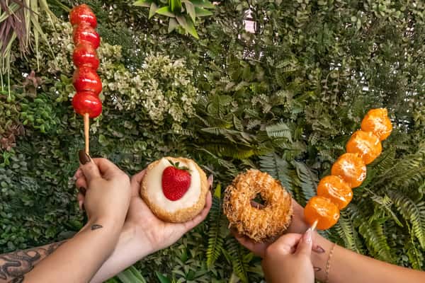 Three hands showcasing Mama Chai's signature treats: glossy candied strawberry and mandarin skewers, a coconut-crusted donut, and a cream-filled strawberry donut, all held in front of a leafy green backdrop.