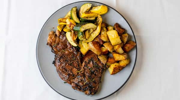 Grilled steak with rosemary, roasted zucchini and yellow squash, and crispy diced potatoes on a gray plate.