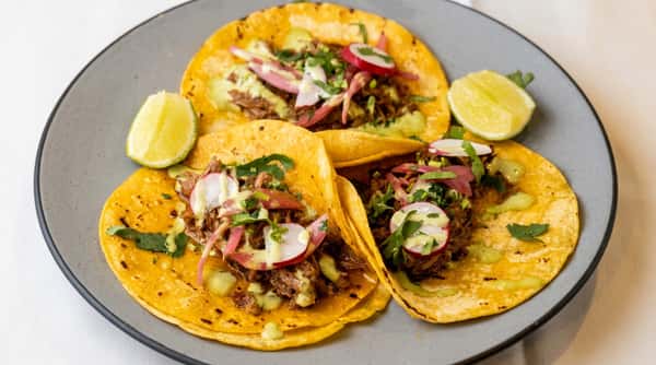 Three street tacos with shredded meat, pickled onions, radishes, cilantro, and green sauce, served with lime wedges.