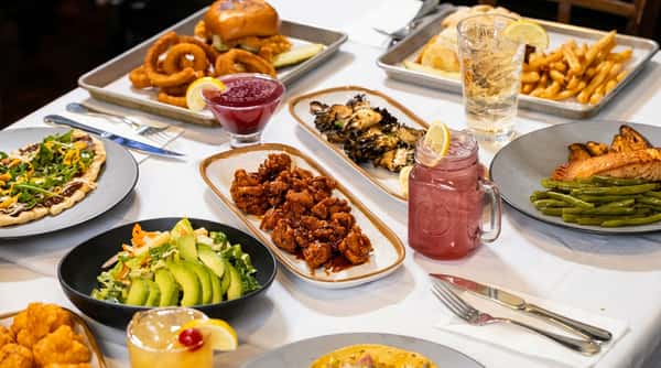 A table laden with multiple dishes including a chicken sandwich, fries, salmon with green beans, tacos, a flatbread, various drinks, and salads.