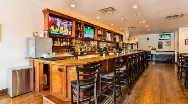 Long wooden bar with black stools, multiple flat-screen TVs, and liquor bottles on shelves, viewed from seating area.