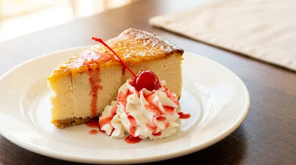 Slice of cheesecake with caramel topping, strawberry drizzle, whipped cream, and a maraschino cherry.