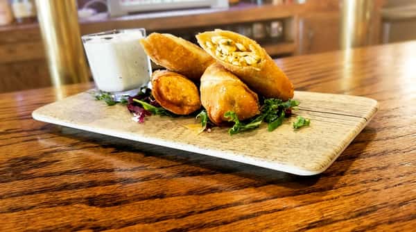 Golden-brown egg rolls with a visible chicken and corn filling, served with a side of ranch dressing and greens