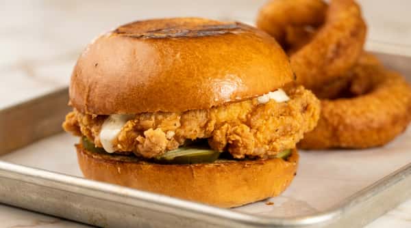 Fried chicken sandwich with pickles and white sauce on a toasted bun, served on a metal tray.