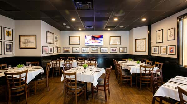 A restaurant dining room with white tablecloths, wooden chairs, dark wood floors, white walls, and a black ceiling.