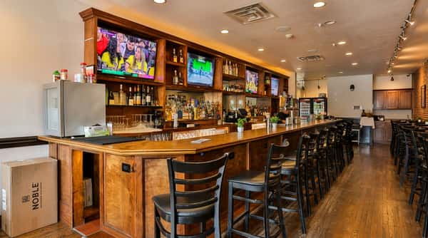 L-shaped wooden bar with black stools, multiple TVs showing sports, and shelves of liquor bottles.