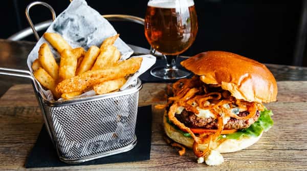 A burger with blue cheese, crispy onions, lettuce, and tomato, alongside fries in a metal basket and a glass of beer.
