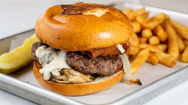 A burger on a brioche bun with a beef patty, grilled mushrooms, onions, melted white cheese sauce, and a side of fries and a pickle spear.