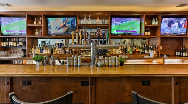 Brightly lit wooden bar with multiple TV screens showing sports, beer taps, and shelves of liquor bottles.