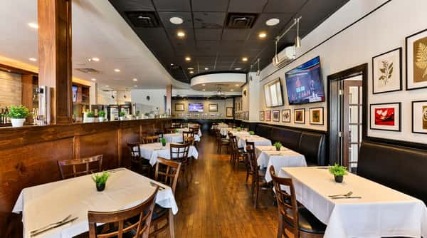 Restaurant dining room with white tablecloths, wooden chairs, dark booths, and TVs on white walls.