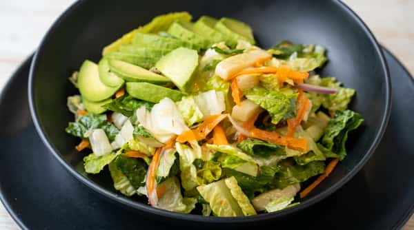 Green salad with sliced avocado, shredded carrots, iceberg lettuce, and red onion in a dark bowl.