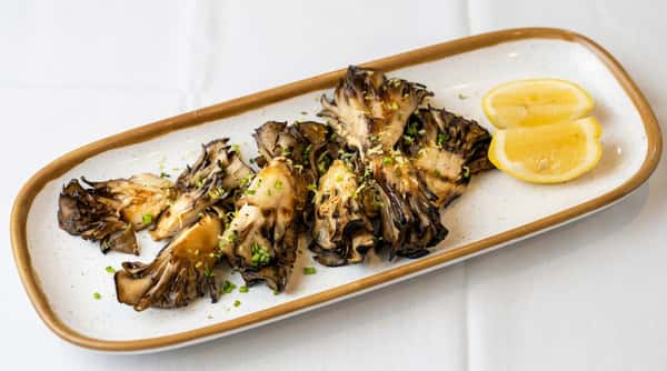 Grilled maitake mushrooms with lemon zest, chives, and two lemon wedges on a white rectangular plate.