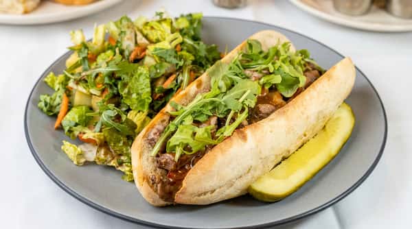 Philly cheesesteak sandwich topped with arugula, served with a side salad and a pickle spear on a gray plate.