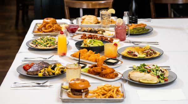 Table laden with various dishes: a burger and fries, chicken wings, popcorn chicken, tacos, and multiple cocktails.