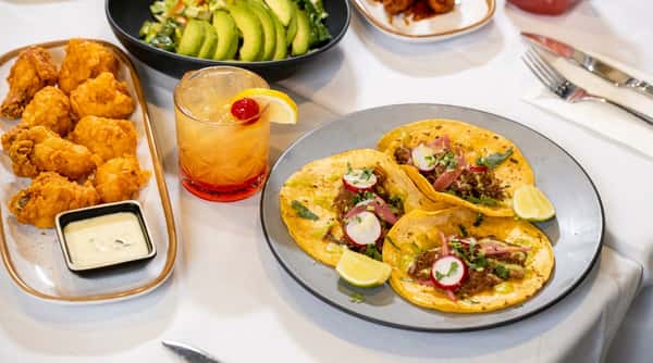 Tacos with meat, radish, cilantro, and lime, beside fried fritters, a colorful cocktail, and a salad.