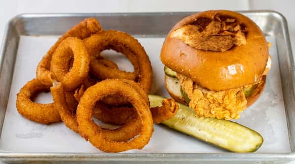 Chicken sandwich with pickles on a brioche bun, served with a pile of onion rings and a pickle spear.