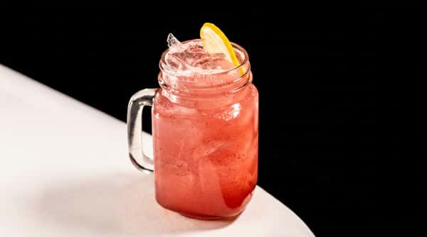 Pink drink in a mason jar mug with ice and a lemon slice, on a white tablecloth.