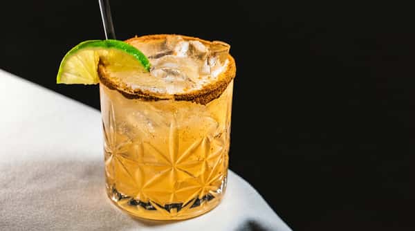 A clear glass with a light-colored cocktail, ice, a dark straw, and a lime wedge. The rim is garnished with a brown spice.