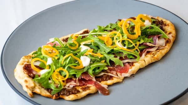 Flatbread with prosciutto, arugula, what appear to be candied lemon peels, and white cheese on a gray plate.