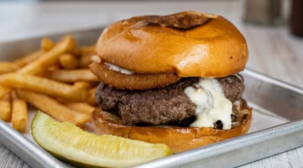 Hamburger with white sauce, onion ring, and a pickle spear on a metal tray.