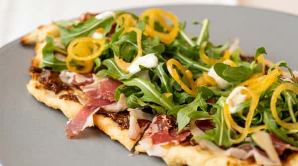 Flatbread or pizza with prosciutto, arugula, golden pepper strips, and white sauce on a gray plate.