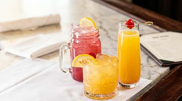 Pink drink with lemon slice in a mason jar, amber drink with lemon and cherry, and a tall flute with orange drink and cherry.