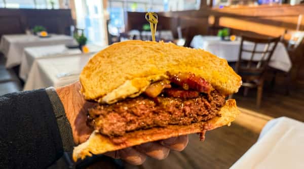Hand holding a thick burger with bacon, cheese, and mustard on a toasted bun, pink meat visible, in a restaurant.