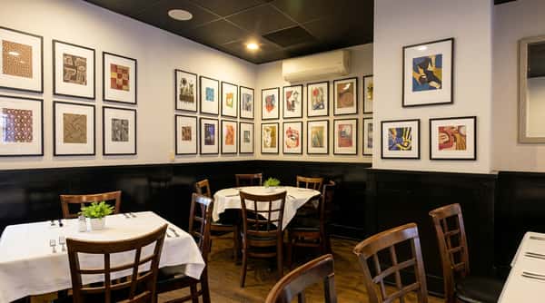 Restaurant dining room with white walls, dark wainscoting, framed artwork, and tables with white tablecloths.