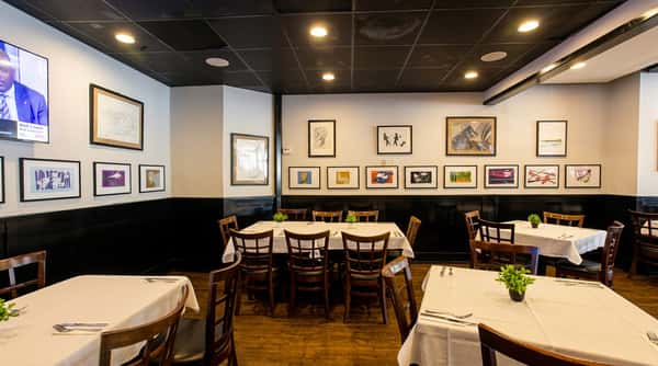 Restaurant dining area with white walls, black wainscoting, a dark ceiling, and various framed art. Tables have white tablecloths and dark wooden chairs.