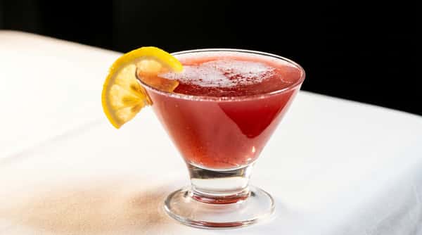 Martini glass filled with a deep red drink, garnished with a yellow lemon slice on a white tablecloth.