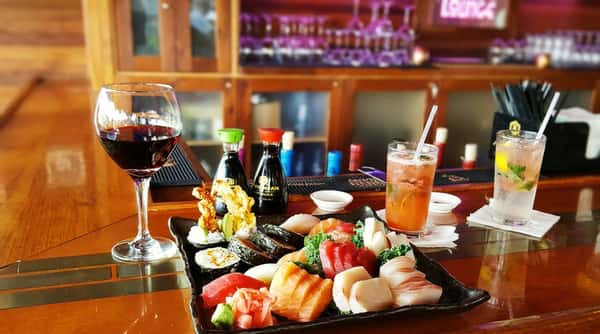 Sushi and sashimi platter with soy sauce, red wine, and a cocktail on a bar counter.