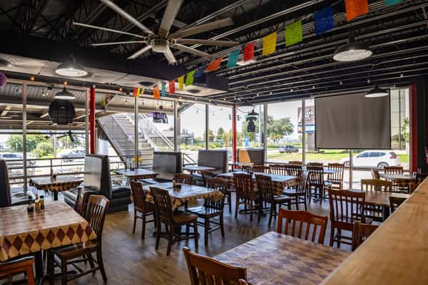 Spacious Mexican restaurant dining area with large windows, papel picado banners, and wooden tables — casual atmosphere with natural light and street-facing views.