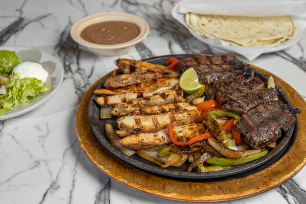 Sizzling mixed fajita platter with grilled chicken and steak strips over sautéed bell peppers and onions, served with refried beans, warm flour tortillas, shredded lettuce, and sour cream — bold Tex-Mex entrée perfect for sharing.