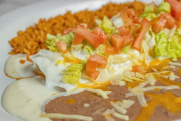 Close-up of cheesy chicken enchiladas topped with fresh lettuce, diced tomatoes, and rich white queso, paired with rice and refried beans.
